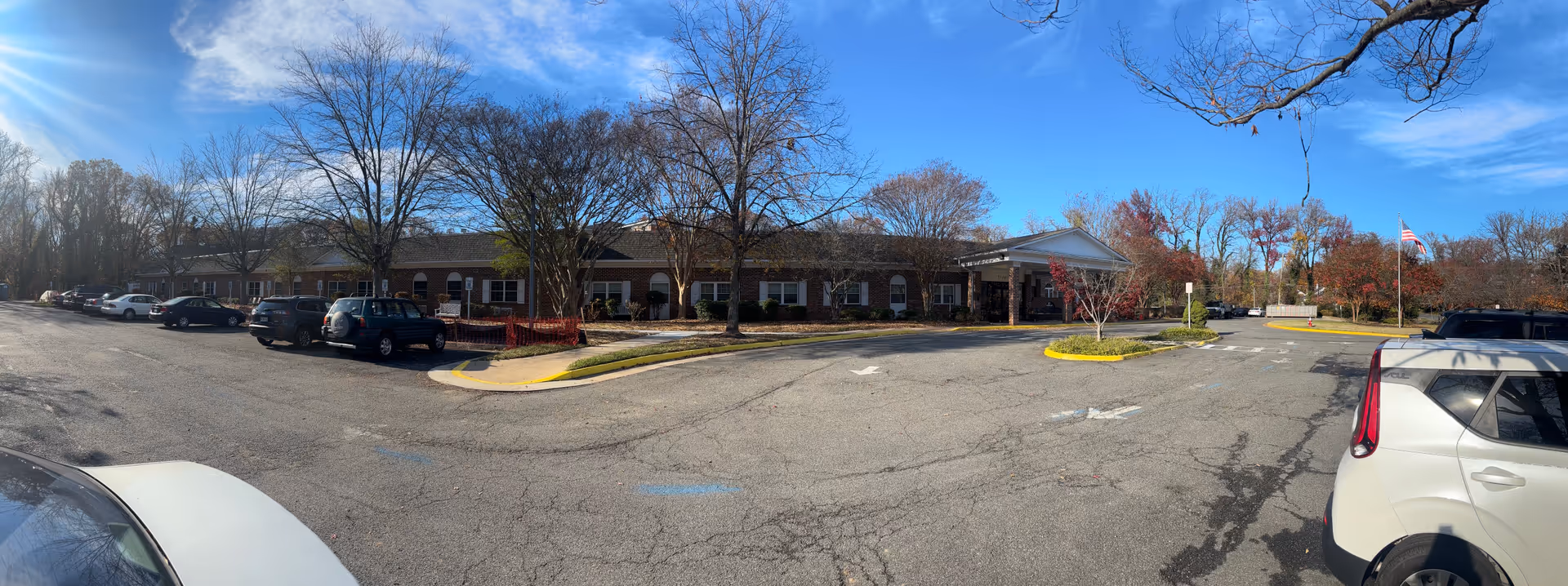 Panoramic view of the front of a single-story brick retirement community building with an entrance canopy, parking lot, trees, and parked cars under a blue sky.