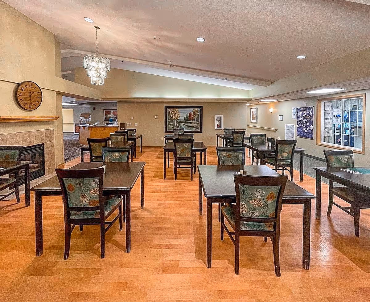 A dining room with several dark wooden tables and chairs with patterned upholstery. The room has a wooden floor, a wall clock above a fireplace on the left, a chandelier hanging from the ceiling, and framed artwork on the walls. There is a window on the right side and a bulletin board with notices.
