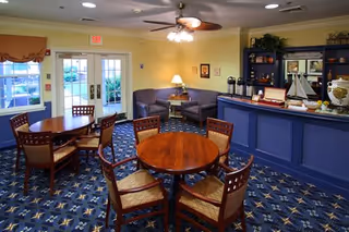 A cozy common area with round wooden tables and chairs on a patterned carpet. There is a blue counter with decorative items including a model sailboat, coffee dispensers, and a lamp. The walls are painted yellow, and there are double glass doors leading outside with a view of greenery.