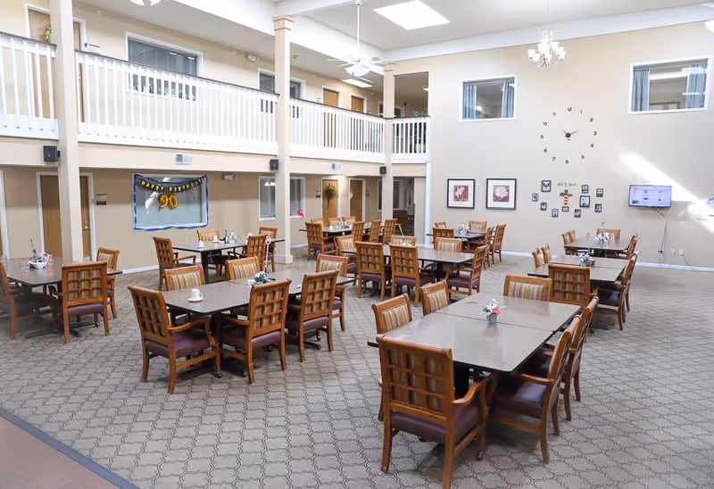 A spacious dining room with multiple wooden tables and chairs arranged neatly. The room has a high ceiling with ceiling fans and a large wall clock. There are framed pictures and a small gallery of photos on the wall. A banner reading 'Happy Birthday 90' is displayed on one side. The room has a carpeted floor and a balcony railing overlooking the area.