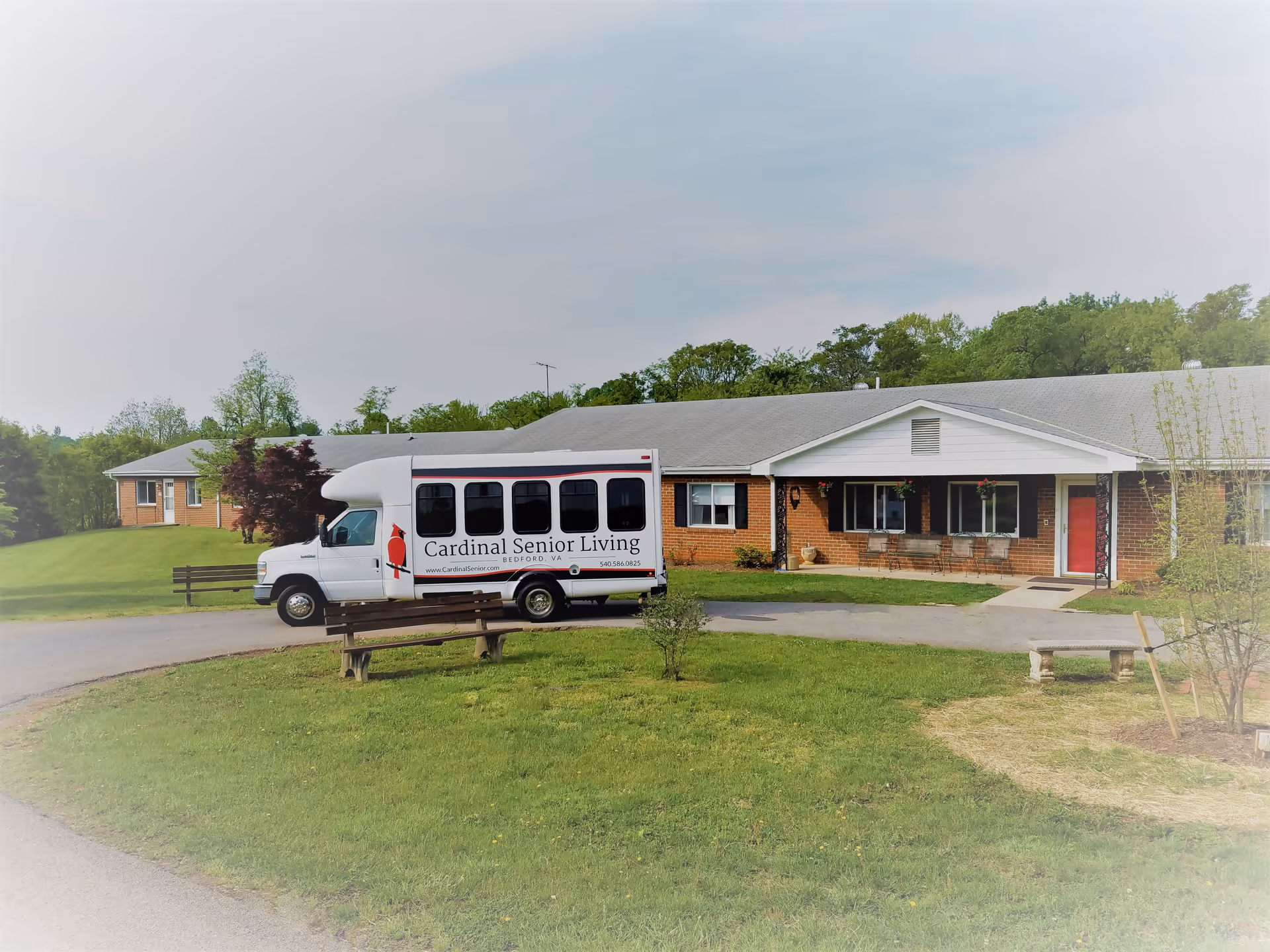 Exterior view of a single-story brick building with a gray roof and a red door, surrounded by green grass and trees. A white shuttle bus with the text 'Cardinal Senior Living Bedford, VA' is parked in front of the building. There are wooden benches on the grass near the driveway.