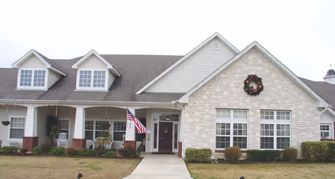Front exterior of a single-story brick and siding senior living building with a covered porch, American flag, wreath, and walkway.