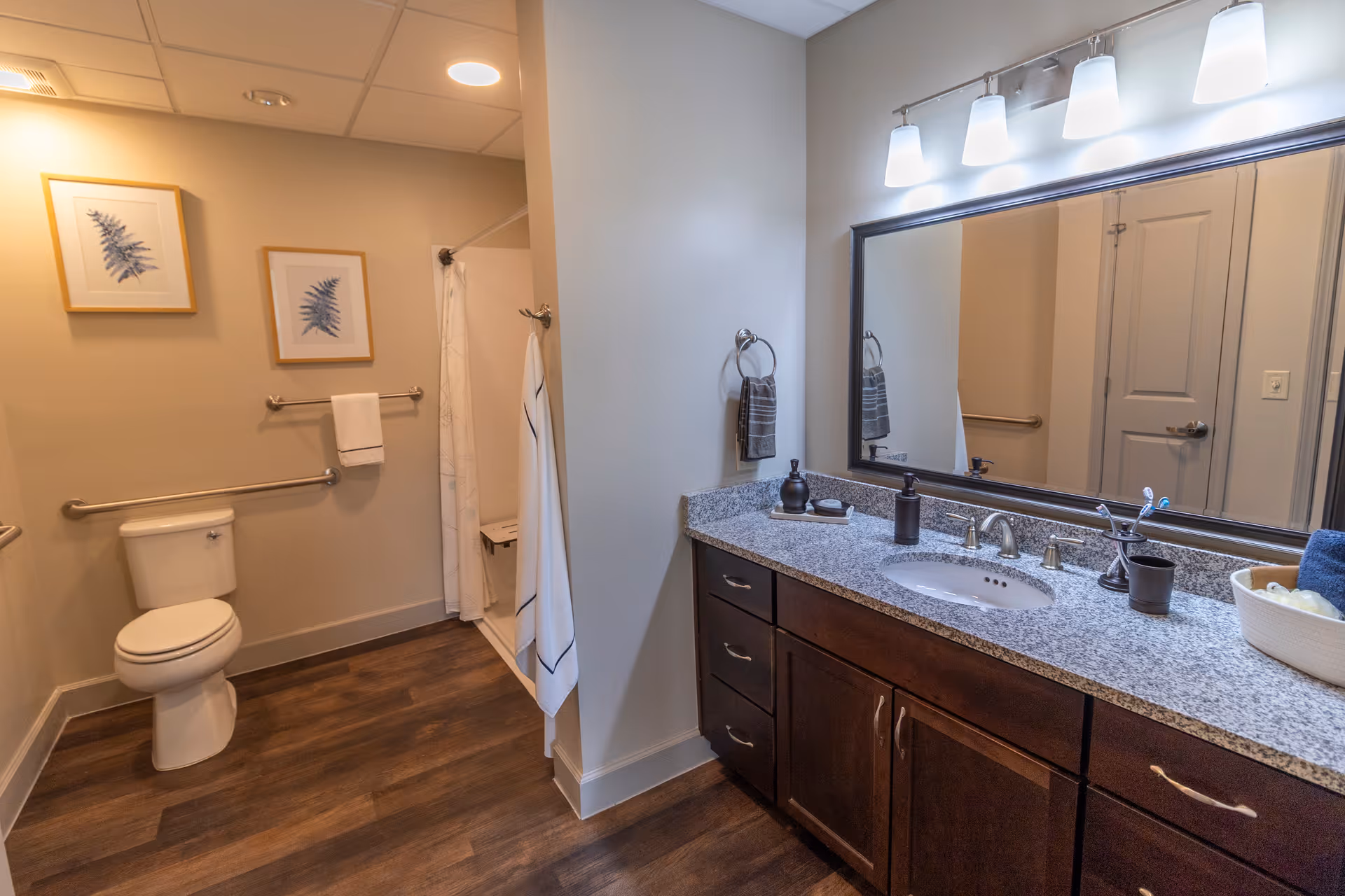 A clean and modern bathroom with a toilet, a walk-in shower with a white curtain, and a large vanity with a granite countertop and a sink. Above the vanity is a large mirror with four lights. The walls are light-colored, and there are two framed botanical prints above the toilet. The floor has wood-style flooring.