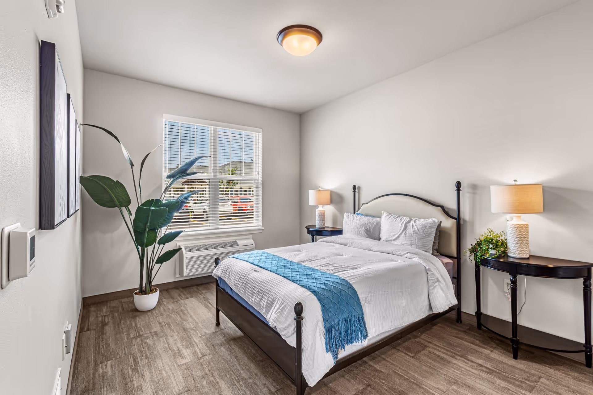 A bright and clean bedroom with a large window letting in natural light. The room features a neatly made bed with white bedding and a blue throw blanket, two bedside tables each with a lamp, a potted plant near the window, and light-colored walls and flooring.