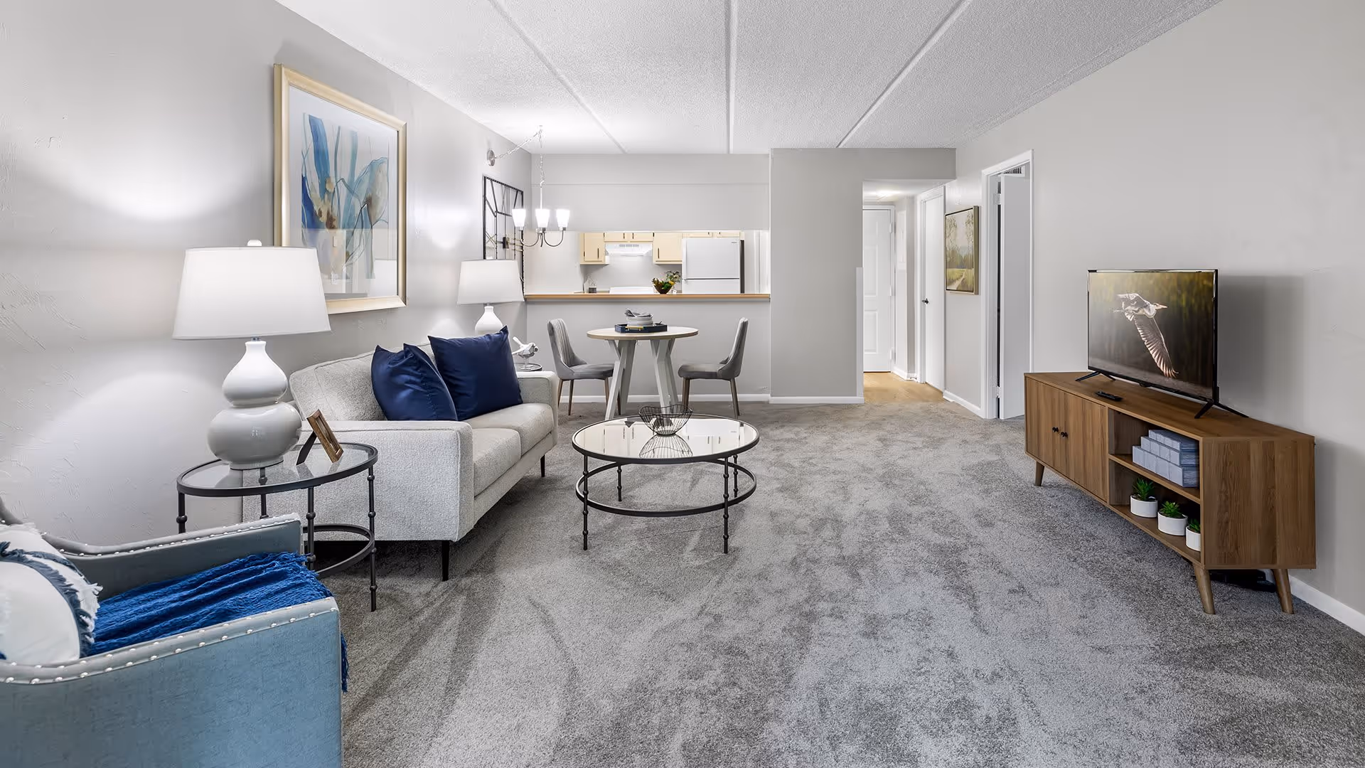 A modern living room with gray carpet, a light gray sofa with dark blue pillows, a blue armchair with a white pillow and blue throw, two white lamps on glass side tables, a round glass coffee table, a small round dining table with two gray chairs, and a wooden TV stand with a flat-screen TV displaying a bird in flight. The kitchen area is visible in the background with white cabinets and appliances.