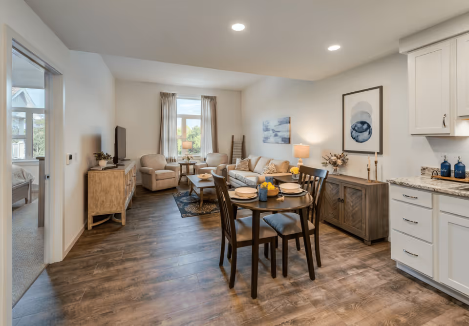 A bright and cozy living and dining area in a senior living facility with a round dining table set for four, a beige sofa, two armchairs, a wooden TV stand with a flat-screen TV, and a sideboard with decorative items. The room has wood flooring, neutral-colored walls, and a window with curtains letting in natural light. A glimpse of a bedroom is visible through an open doorway on the left.