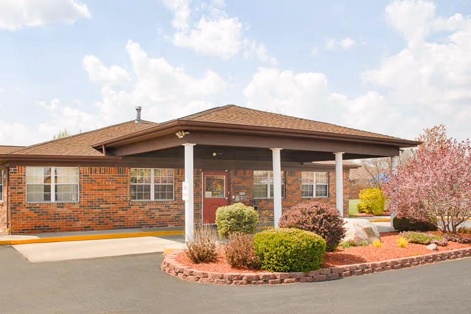 Exterior view of a single-story brick building with a covered entrance supported by white columns. The building has multiple windows and a red door. There are landscaped bushes and small trees with red mulch around the entrance area under a partly cloudy sky.