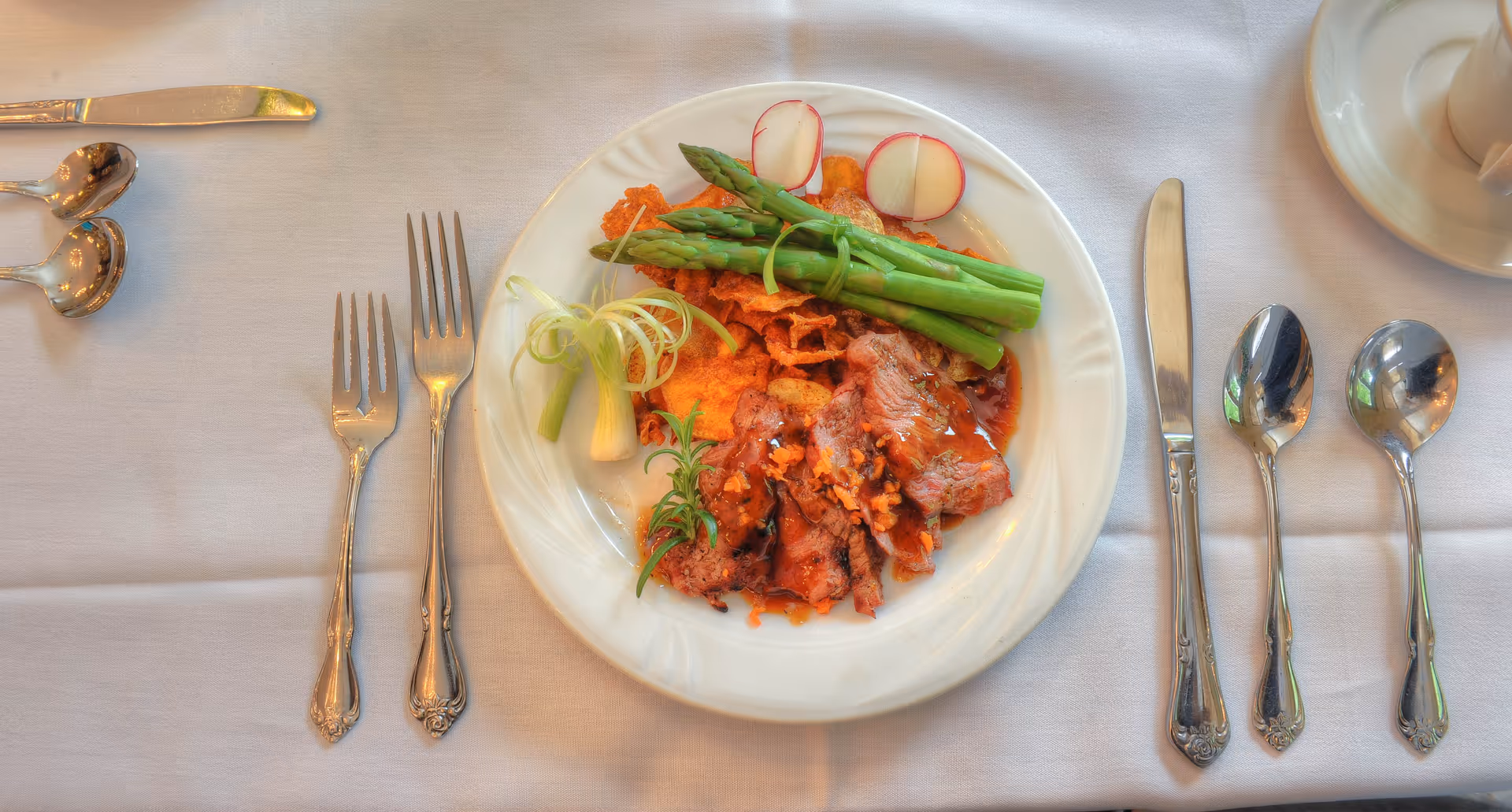 A white plate with a meal consisting of sliced meat with sauce, asparagus, radish slices, and garnishes, placed on a white tablecloth. Surrounding the plate are two forks on the left and a knife and two spoons on the right, along with a cup and saucer in the top right corner.