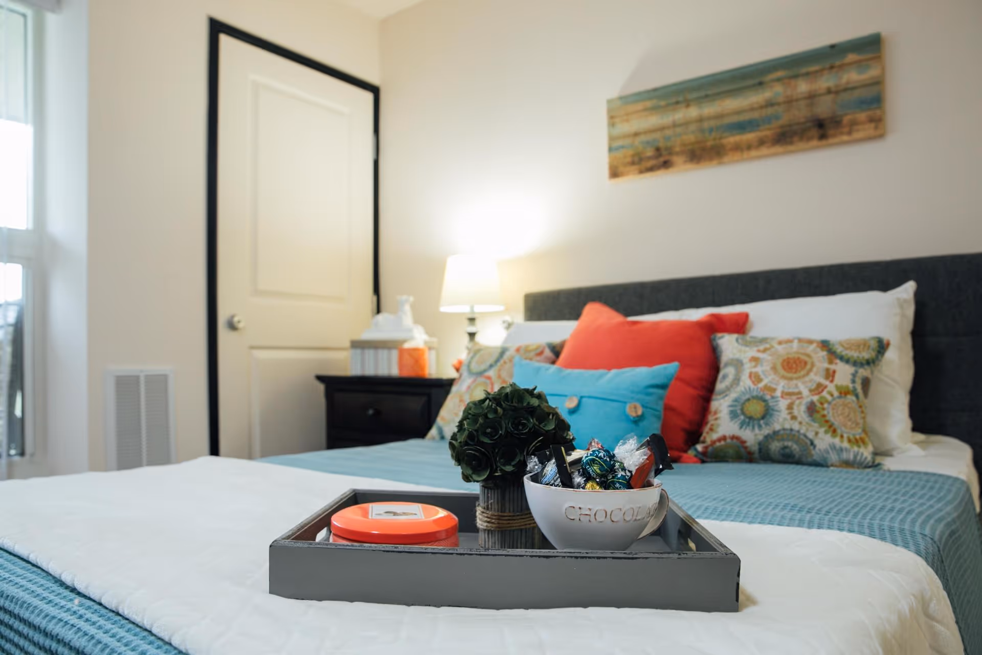 A neatly made bed with a gray headboard, colorful pillows including red, blue, and patterned ones, and a blue blanket. On the bed is a tray holding a small potted plant, a bowl labeled 'CHOCOLATE' filled with assorted candies, and a round orange container. In the background, there is a nightstand with a lamp, tissue box, and a closed white door. A window is partially visible on the left side of the image, and a piece of artwork hangs on the wall above the bed.