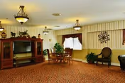 A spacious common area with patterned carpet, a wooden entertainment center with a television, a round table with chairs, decorative plants, and wall decorations including a clock and framed window curtains.