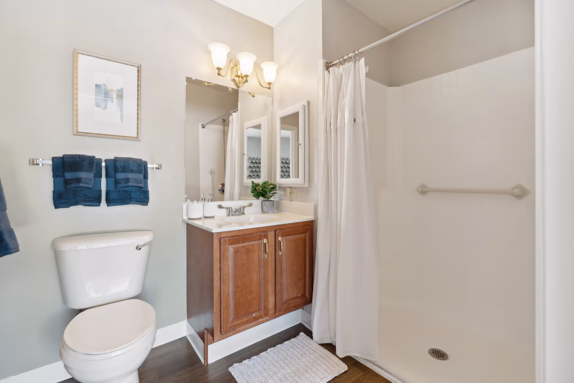 A clean and well-lit bathroom featuring a white toilet, a wooden vanity with a white countertop and sink, a large mirror above the sink, a wall-mounted light fixture with three bulbs, a white shower area with a curtain, and two dark blue towels hanging on a towel rack. There is also a small plant and some toiletries on the vanity.