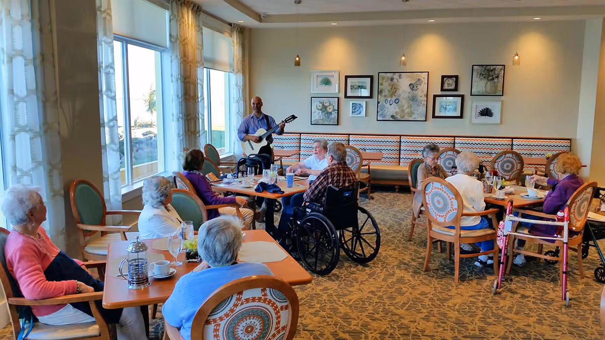 A group of elderly people sitting around tables in a dining room, some in wheelchairs and others with walkers, while a man stands playing a guitar near the windows. The room has large windows with curtains, patterned chairs, and framed artwork on the wall.