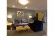 A cozy living room area in a senior living facility featuring a patterned sofa, a dark armchair, a wooden coffee table with a small plant, two side tables each with a lamp, a framed picture on the wall, and a doorway leading to another room.
