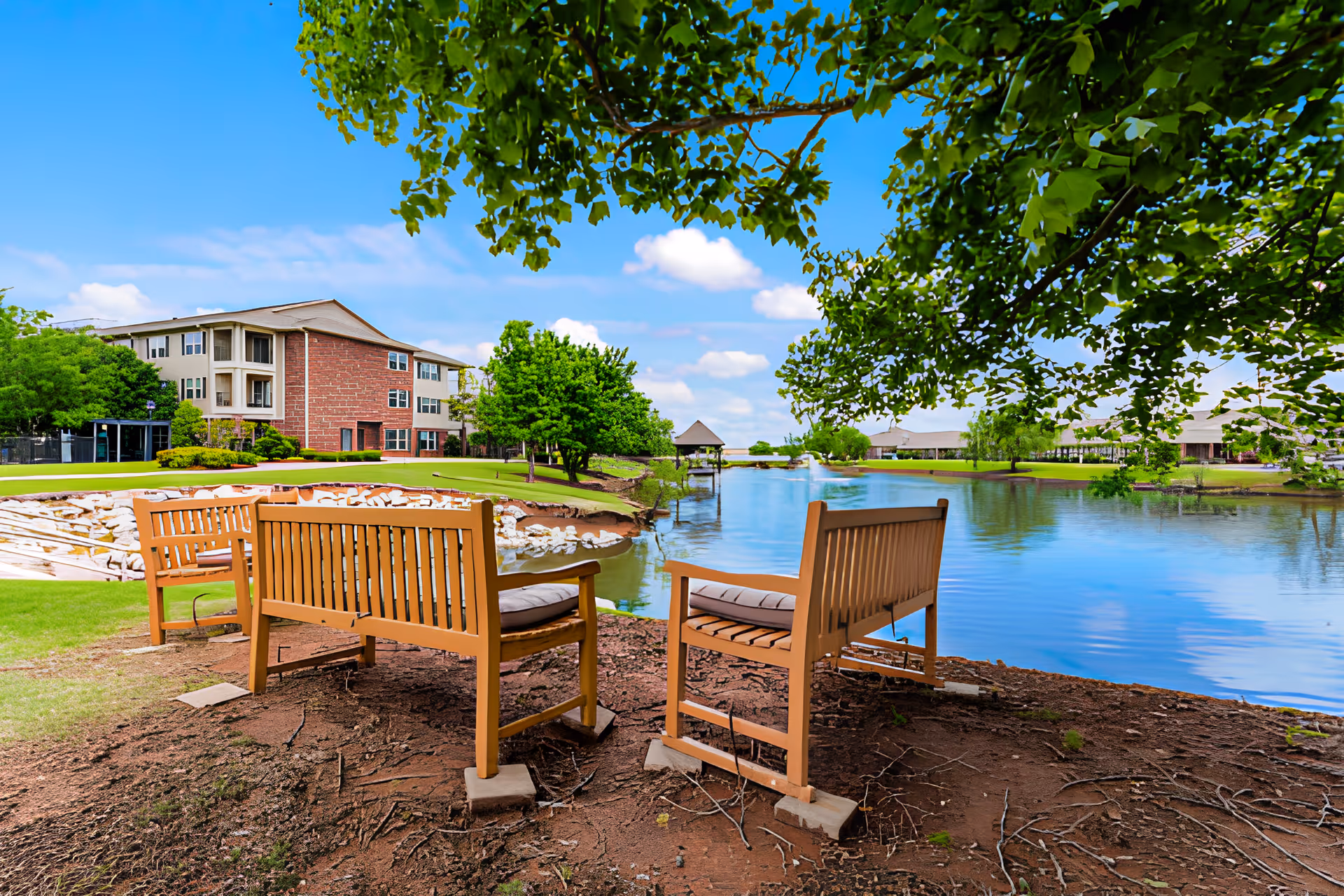 Three wooden benches with cushions arranged near the edge of a calm pond surrounded by green grass and trees, with a multi-story brick and beige building in the background under a blue sky with scattered clouds.
