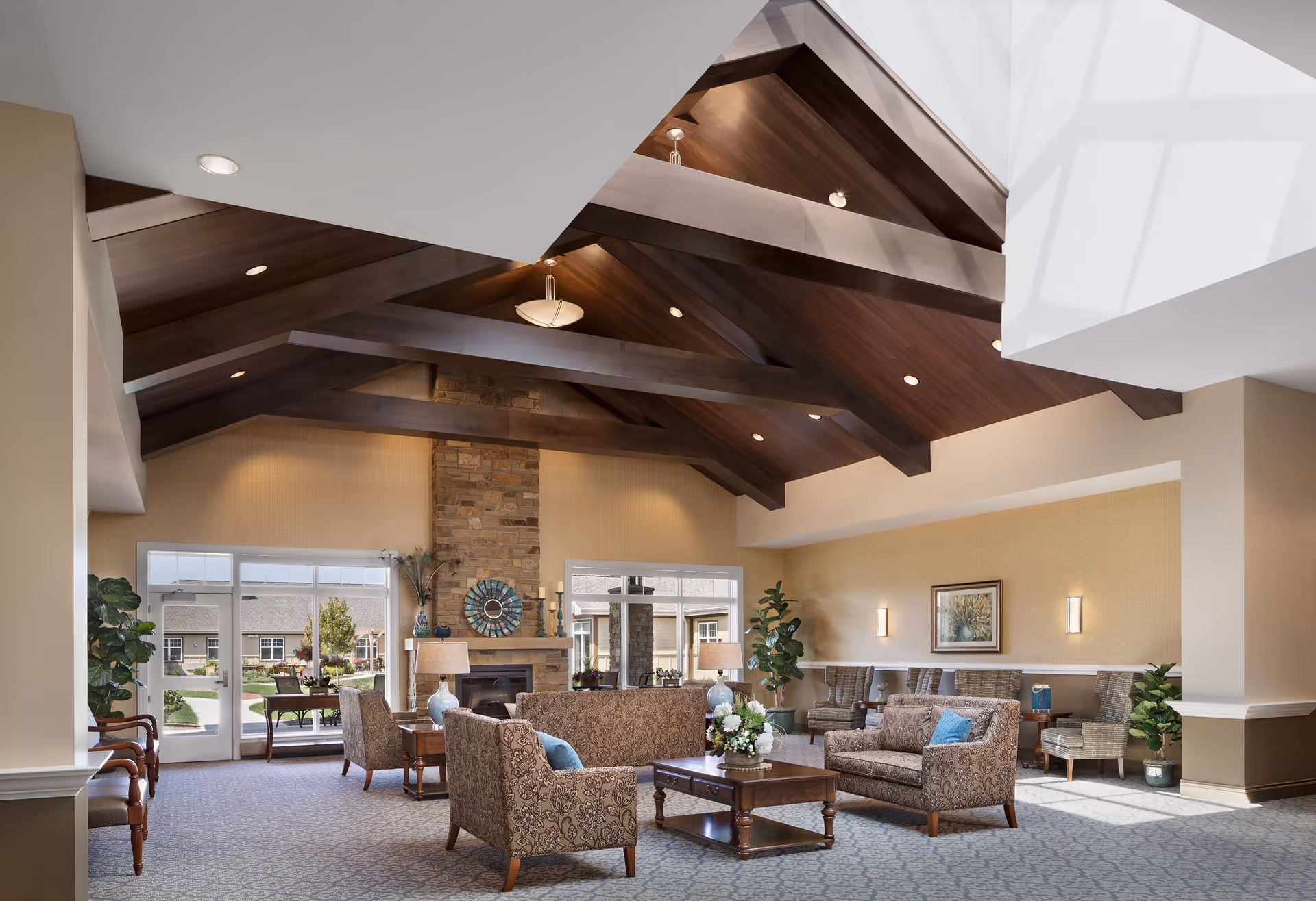 A spacious and well-lit living room area in Silverado St. Charles featuring a high vaulted ceiling with exposed wooden beams and recessed lighting. The room has a stone fireplace centered between two large windows and glass doors that lead outside. Comfortable patterned sofas and armchairs with blue accent pillows are arranged around a wooden coffee table with a floral centerpiece. Additional seating is placed along the walls, complemented by potted plants and soft wall lighting. The carpet has a subtle geometric pattern, and natural light fills the room through the windows and skylight.