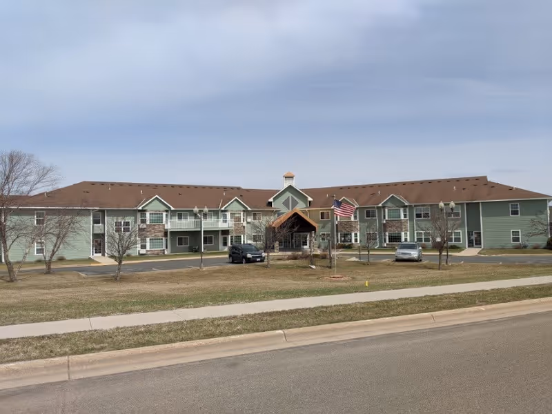 Exterior view of a two-story senior living facility building with green siding and a brown roof. There is a covered entrance with an American flag in front, a few parked cars, leafless trees, and a sidewalk along the street.