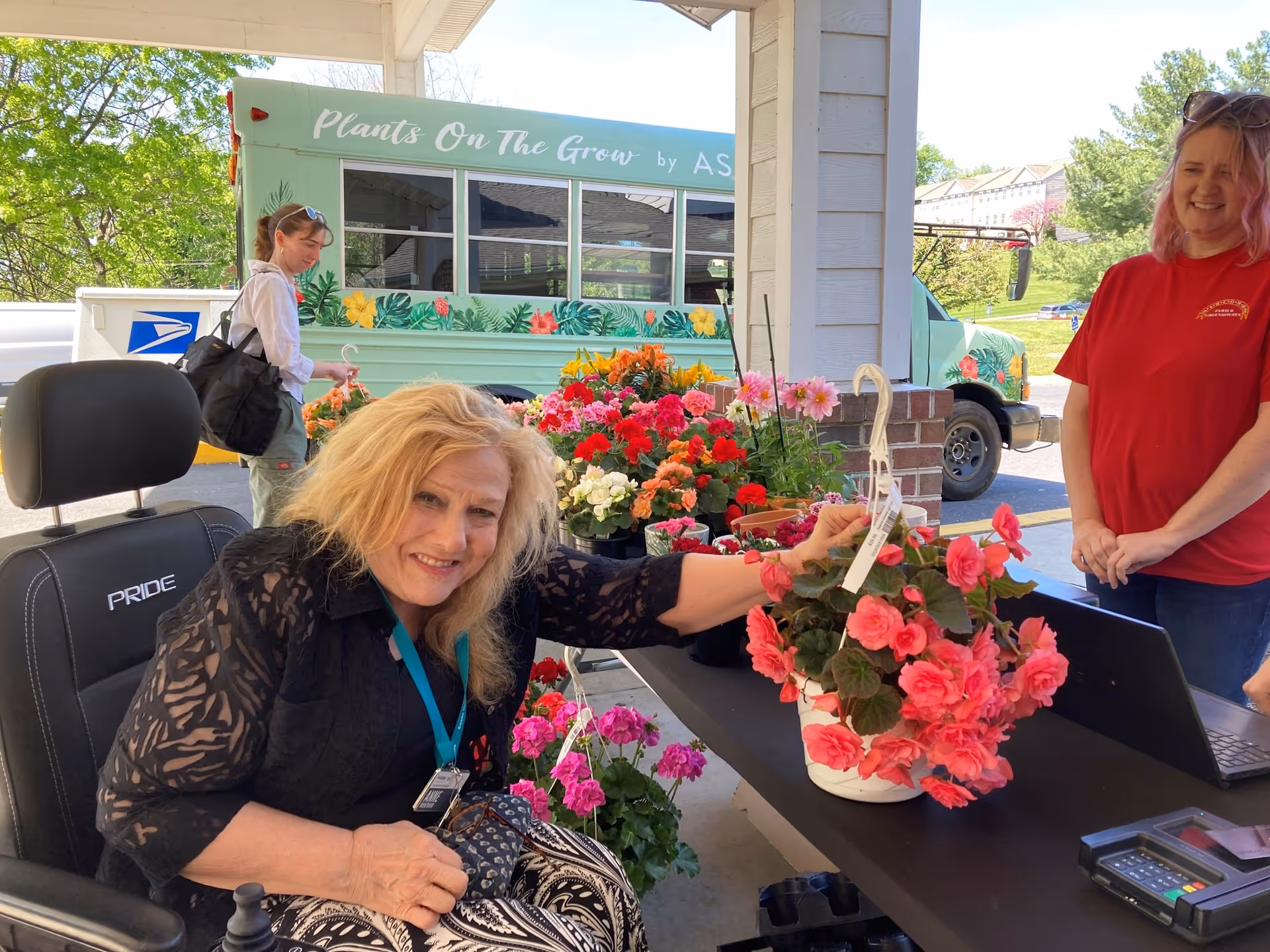A woman in a wheelchair smiling and reaching for a pink flowering plant on a table filled with various colorful flowers. Another woman in a red shirt stands behind the table smiling. In the background, a woman with a backpack is walking past a green bus with floral decorations and the text 'Plants On The Grow'.