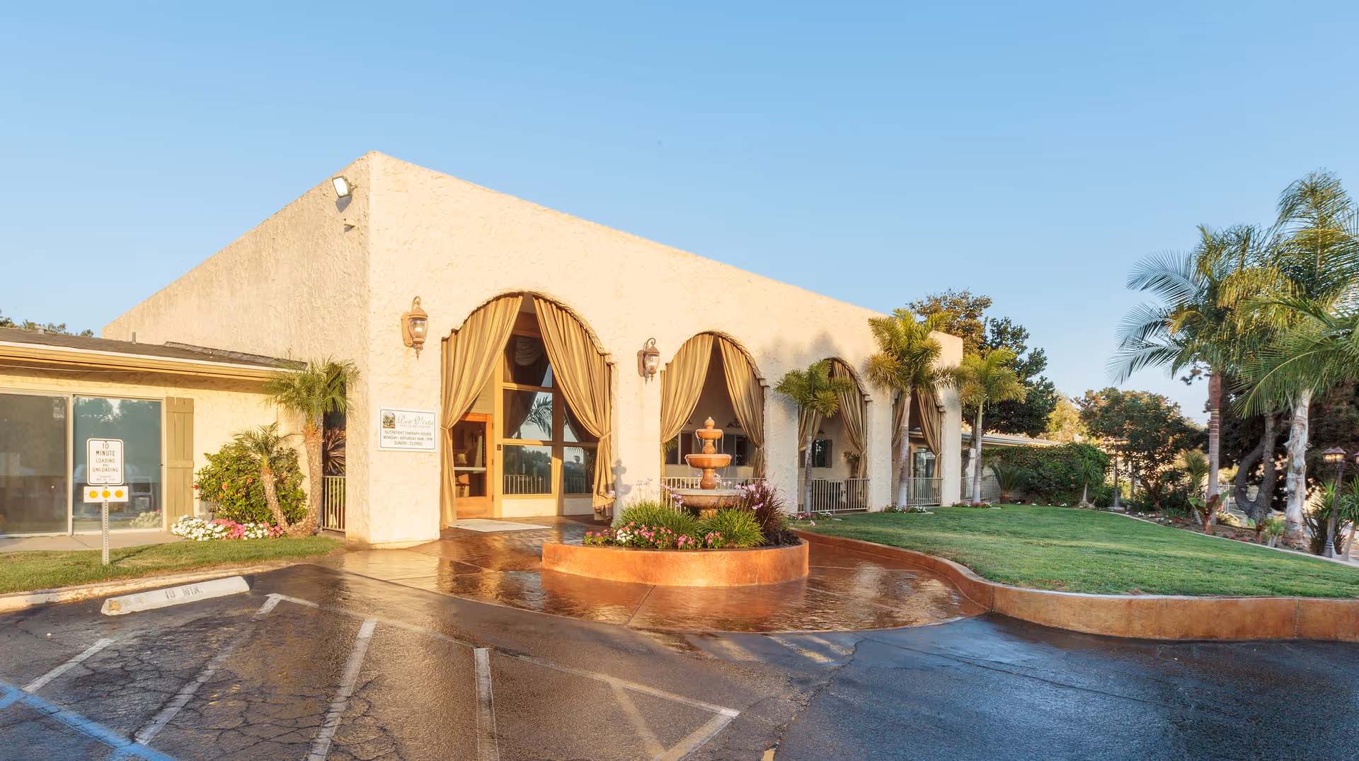 Exterior view of Reo Vista Healthcare Center building with beige stucco walls, arched windows with curtains, a central water fountain surrounded by plants, palm trees, and a parking area in front.