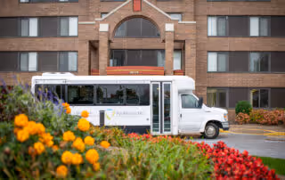 A white shuttle bus parked in front of a multi-story brick building with several windows. In the foreground, there are colorful flowers including yellow and red blooms. The building has an arched window above the entrance.