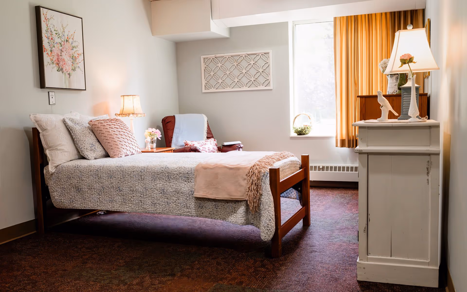 A cozy bedroom in an assisted living facility featuring a single wooden bed with a gray and pink quilt, multiple pillows, a pink throw blanket, a bedside table with a lamp and flowers, a comfortable armchair with a blanket, a window with orange curtains, and a white dresser with a lamp and decorative items.