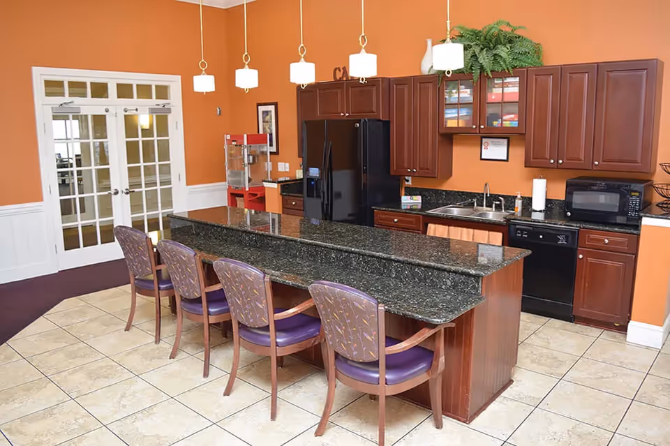 A kitchen area with a long granite countertop island surrounded by four wooden chairs with purple cushions. The kitchen features dark wood cabinets, a black refrigerator, dishwasher, microwave, and a sink. The walls are painted orange, and there are four pendant lights hanging above the island. In the background, there are double glass doors and a popcorn machine on a small counter.