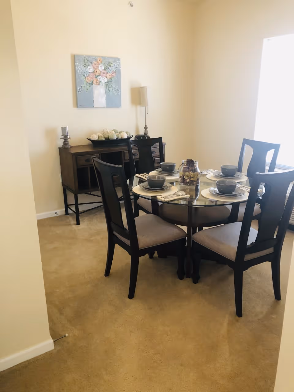 A dining room with a round glass table set for four with matching dark wood chairs with light cushions. On the table are four place settings with bowls, plates, and napkins, and a decorative jar centerpiece. Against the wall is a wooden sideboard with decorative items including a bowl of ornamental balls and a lamp. A floral painting hangs above the sideboard. The room has beige carpet and light-colored walls with a large window letting in natural light.
