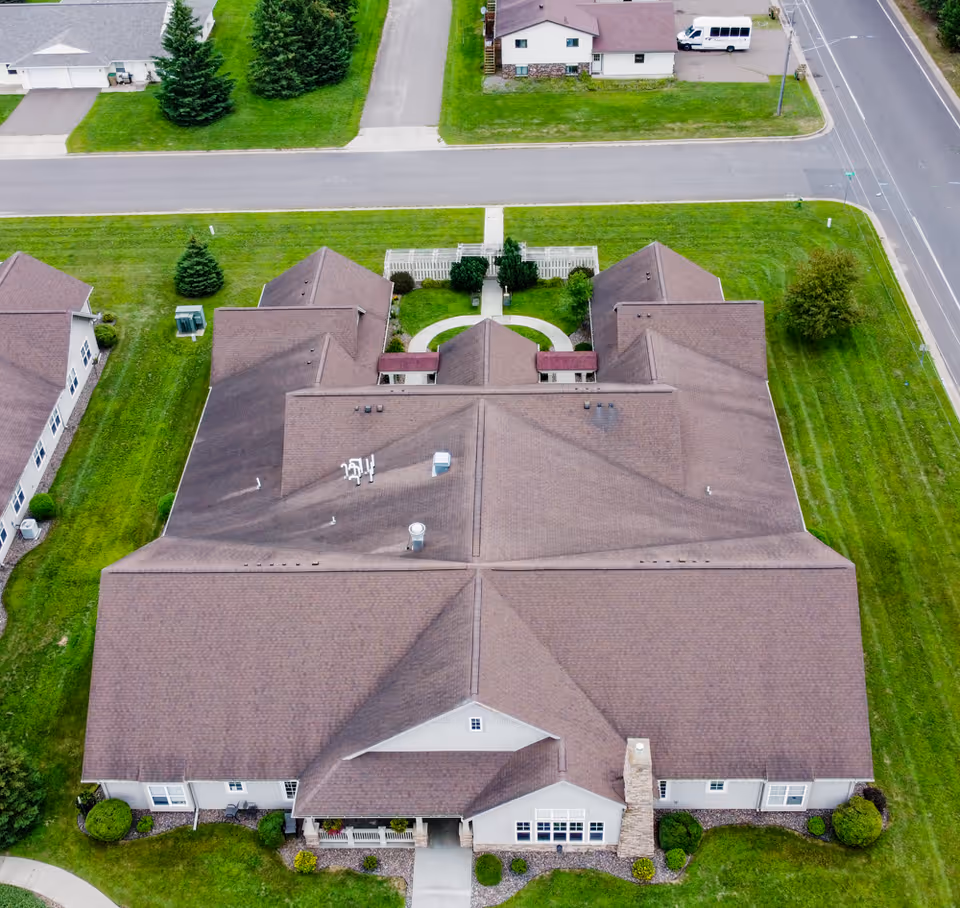 Aerial view of a large single-story building with a brown roof, surrounded by well-maintained green lawns and landscaping. The building has multiple sections and a small courtyard area with a white fence. Nearby roads and neighboring buildings are visible.