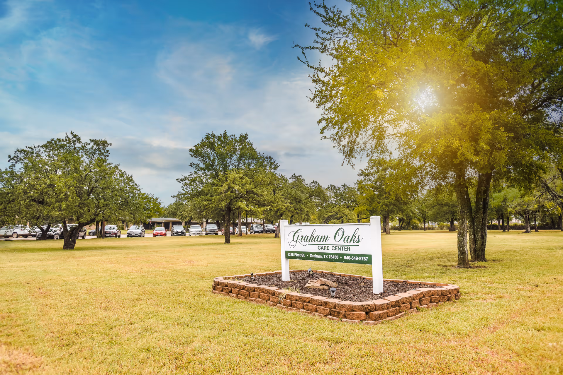 Outdoor view of a grassy area with several trees and a sign that reads 'Graham Oaks Care Center' along with the address and phone number. Cars are parked in the background under a partly cloudy sky with sunlight filtering through the trees.