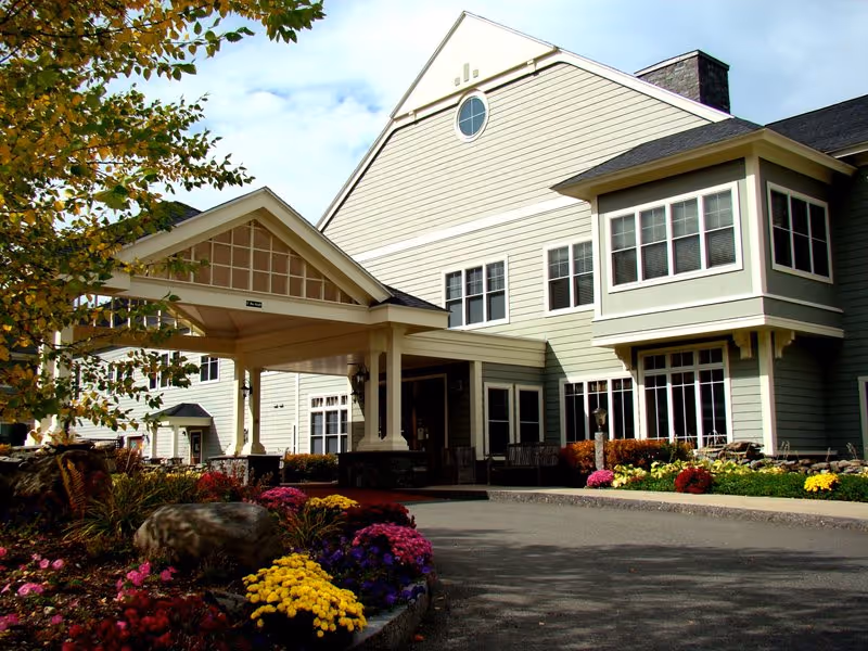 Front exterior of a multi-story senior living building with a covered entrance and landscaped flower beds.