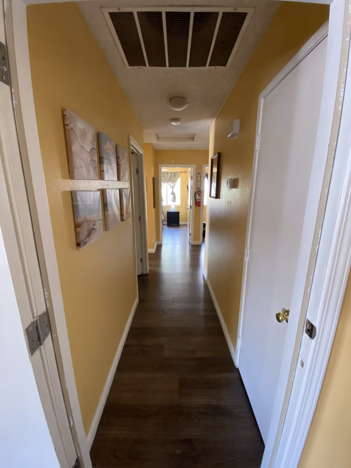 A narrow hallway with yellow walls and dark wood flooring. There are several doors along the hallway, artwork on the left wall, and a window with curtains at the far end letting in natural light.