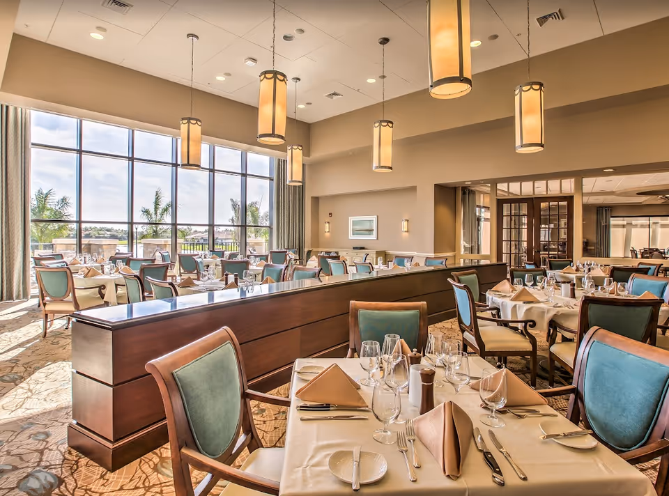 A bright and elegant dining room with large floor-to-ceiling windows letting in natural light. The room features multiple tables set with white tablecloths, beige napkins, glassware, and silverware. The chairs have wooden frames with teal upholstery. Several pendant lights hang from the ceiling, and there is a wooden divider separating sections of the dining area. Outside the windows, palm trees and a clear sky are visible.