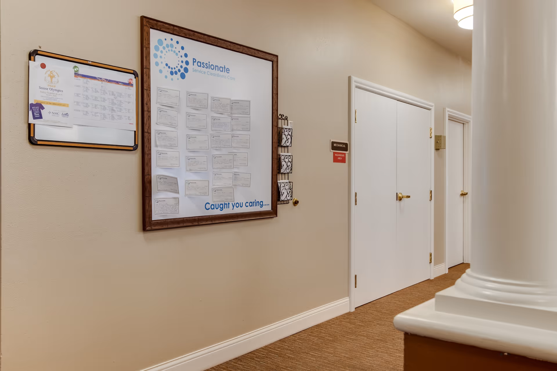 Interior hallway of an assisted living facility with beige walls and carpeted floor. On the left wall, there are two bulletin boards with notices and information, one framed with the heading 'Passionate Service Excellence Care' and the phrase 'Caught you caring...'. On the right side, there are white double doors labeled 'Mechanical' and a smaller door further down the hallway. A white column is partially visible in the foreground on the right.