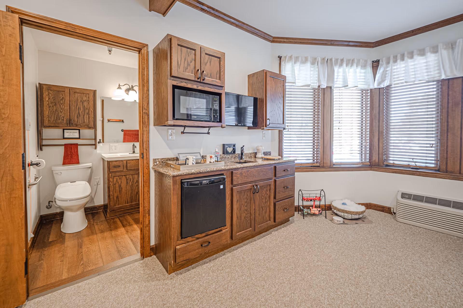 Interior view of a room with wooden cabinetry, a small sink, a microwave, a mini fridge, and a television mounted on the wall. To the left, there is an open door leading to a bathroom with a toilet, a sink with a mirror, and wooden cabinets. The room has carpeted flooring, a bay window with blinds and white curtains, and a pet bed with toys on the floor near the window.