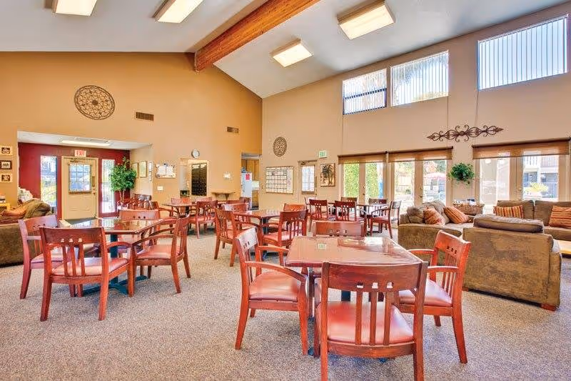 A spacious common area in a senior living facility with multiple wooden tables and chairs arranged for dining or socializing. The room features high ceilings with exposed wooden beams, large windows allowing natural light, and a seating area with sofas and cushions near the windows. The walls are decorated with circular wall art and plants, creating a warm and inviting atmosphere.