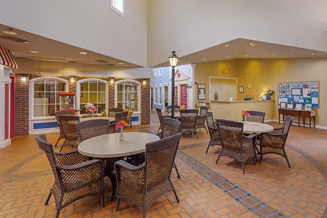 Indoor common area with round tables and wicker chairs arranged on a tiled floor. The space features a high ceiling with recessed lighting, a reception desk in the background, and decorative elements resembling storefront windows and a street lamp, creating a cozy, village-like atmosphere.