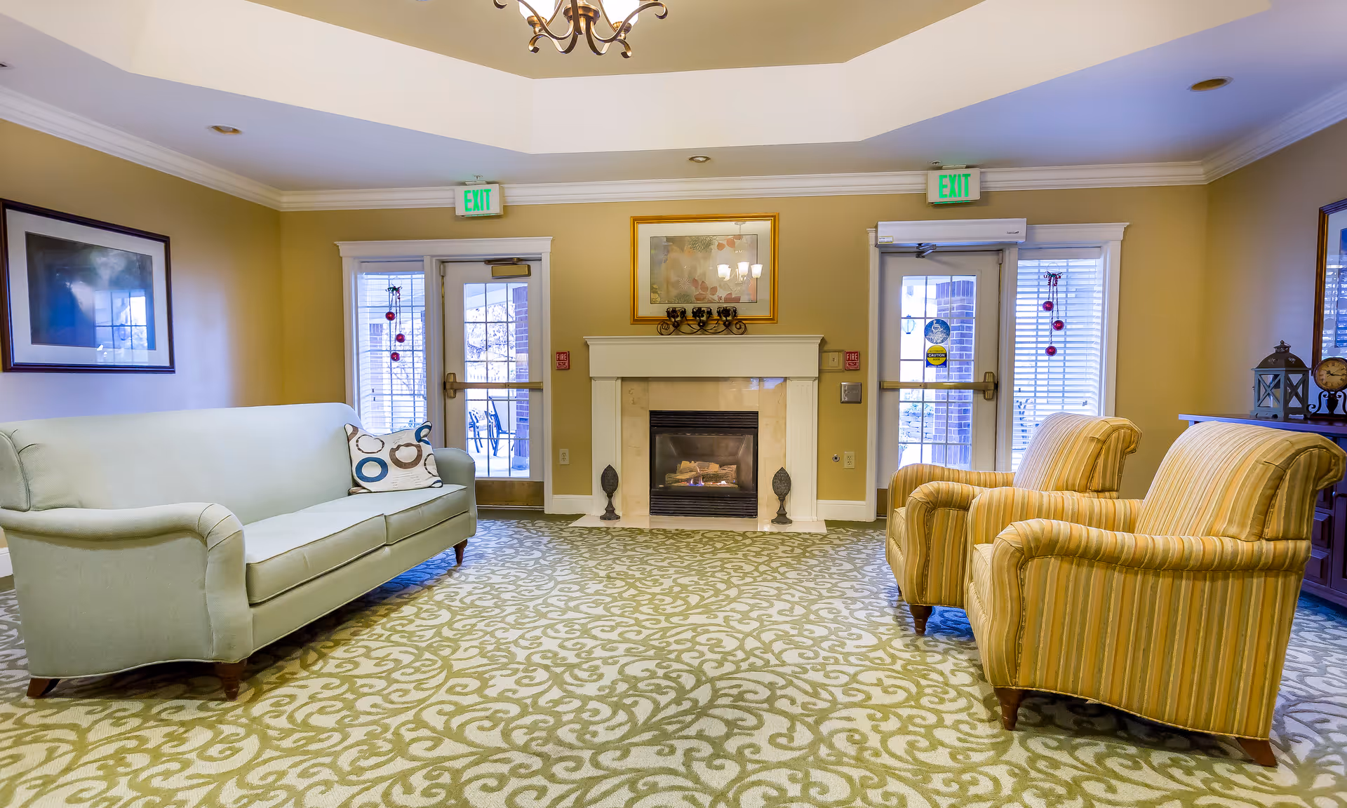 A cozy living room area with a light green sofa on the left and two striped armchairs on the right. The room features a patterned carpet, a fireplace in the center with a framed picture above it, and two glass doors with exit signs above them. The walls are painted beige, and there are decorative items on a side table to the right.