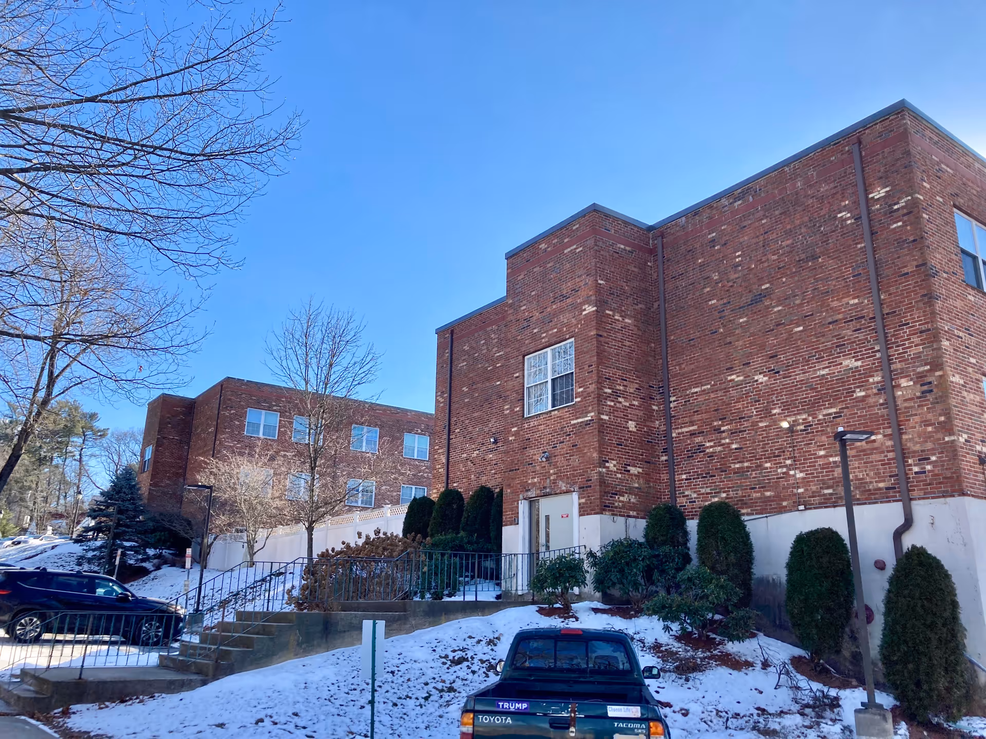 Brick multi-story building exterior with snow on the ground, parked cars, and leafless trees under a clear blue sky.