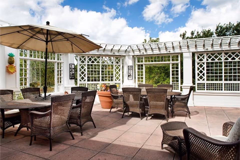 Outdoor patio area with multiple brown wicker tables and chairs, a large beige umbrella providing shade, white lattice fencing, and a large potted plant. The sky is blue with some clouds and greenery is visible beyond the patio.