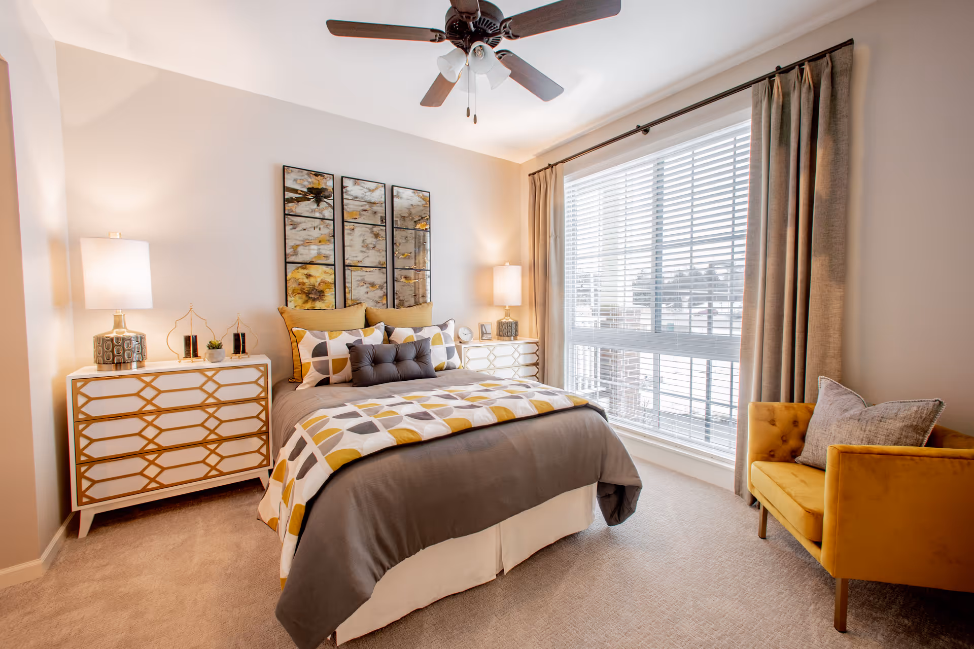 Bright furnished bedroom with a bed, matching nightstands and lamps, decorative wall art, a large window with blinds, and a mustard-yellow armchair.