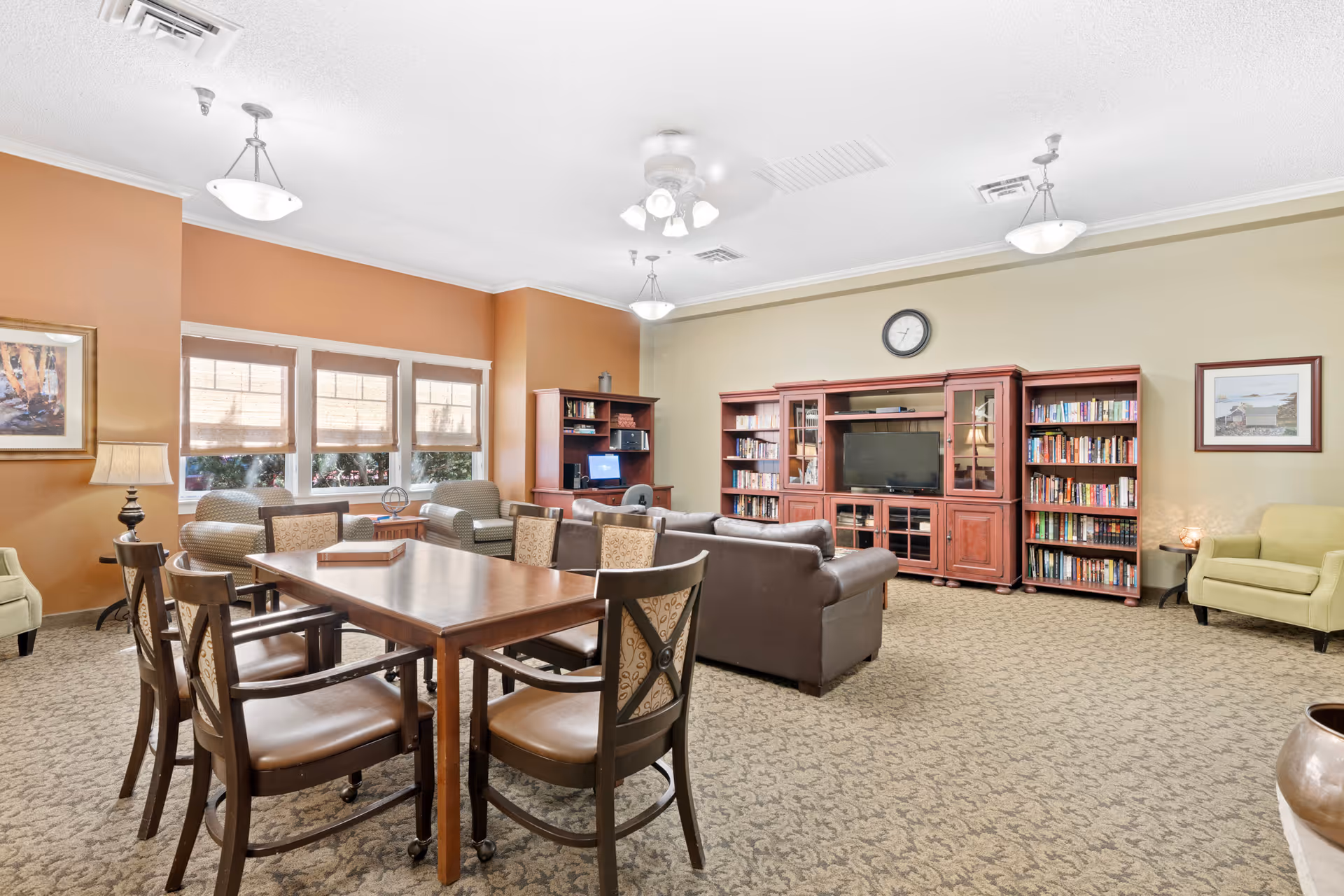 A spacious senior living common area with a wooden table and six chairs in the foreground, two armchairs near a window with blinds, a brown leather sofa facing a large wooden entertainment center with a TV and bookshelves, a computer desk with a chair, and two framed pictures on the walls. The room has beige and light orange walls, carpeted floor, ceiling lights, and a ceiling fan.