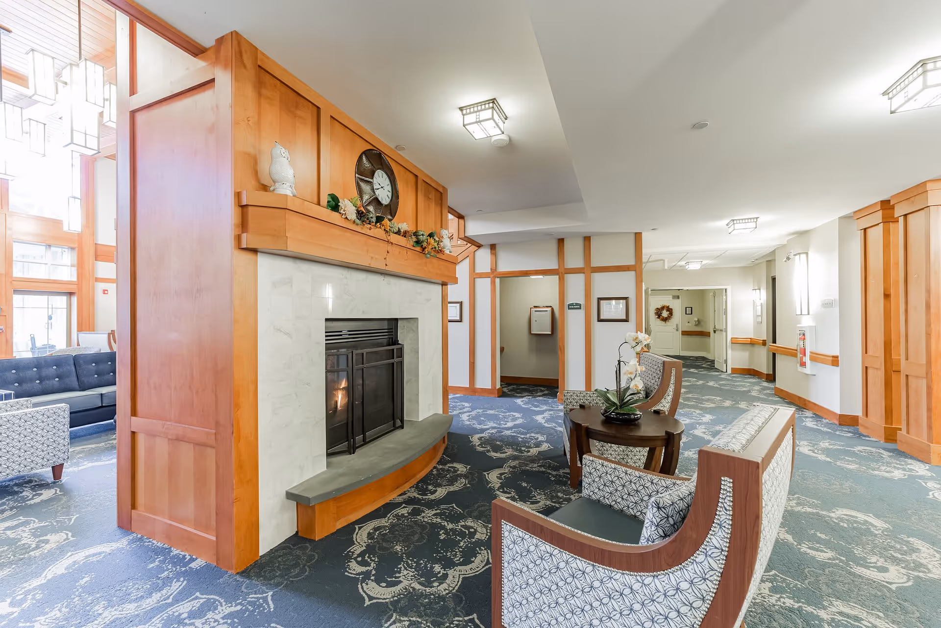 A cozy senior living facility common area with a central fireplace surrounded by wooden paneling. The fireplace has a clock and decorative items on the mantel. There are patterned armchairs and a small round table with a flower arrangement in front of the fireplace. The area has blue patterned carpet and light-colored walls with wooden trim. In the background, there is a hallway leading to other rooms and a seating area with a dark blue couch and a patterned armchair near large windows.