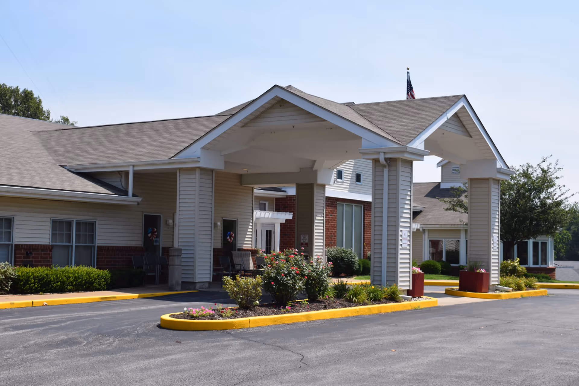 Exterior view of a single-story senior living facility with a covered entrance supported by columns. The building has beige siding with brick accents and a gray shingled roof. There are shrubs and flower beds around the entrance area, and an American flag is visible on the roof.