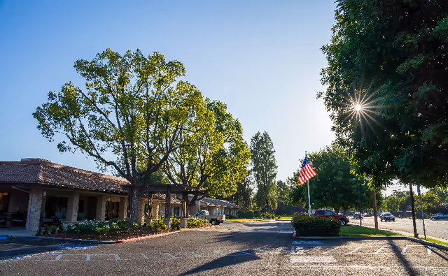 Parking lot area outside a single-story building with a tiled roof, surrounded by large green trees and an American flag on a flagpole. The sun is shining brightly through the trees, casting long shadows on the pavement.