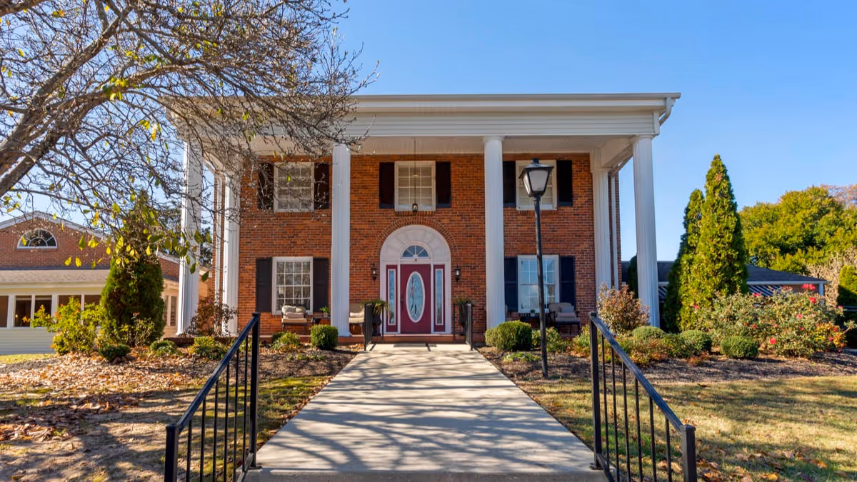 Front view of a two-story brick building with large white columns, a walkway leading to a red double front door, and landscaped grounds.