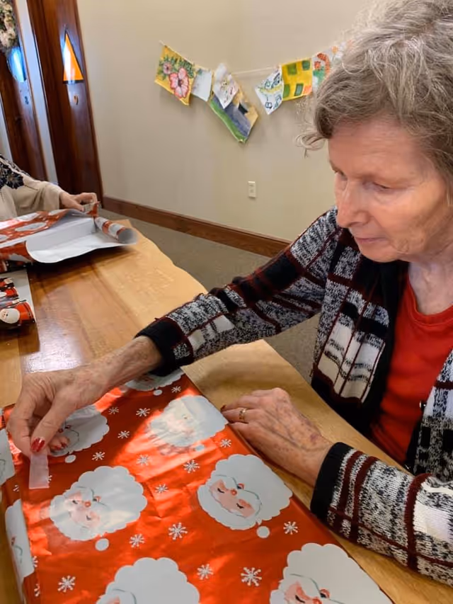 An elderly woman with gray hair is sitting at a wooden table wrapping a gift with red Santa Claus-themed wrapping paper. Another person is partially visible in the background also wrapping a gift. Colorful drawings are hung on the wall behind them.