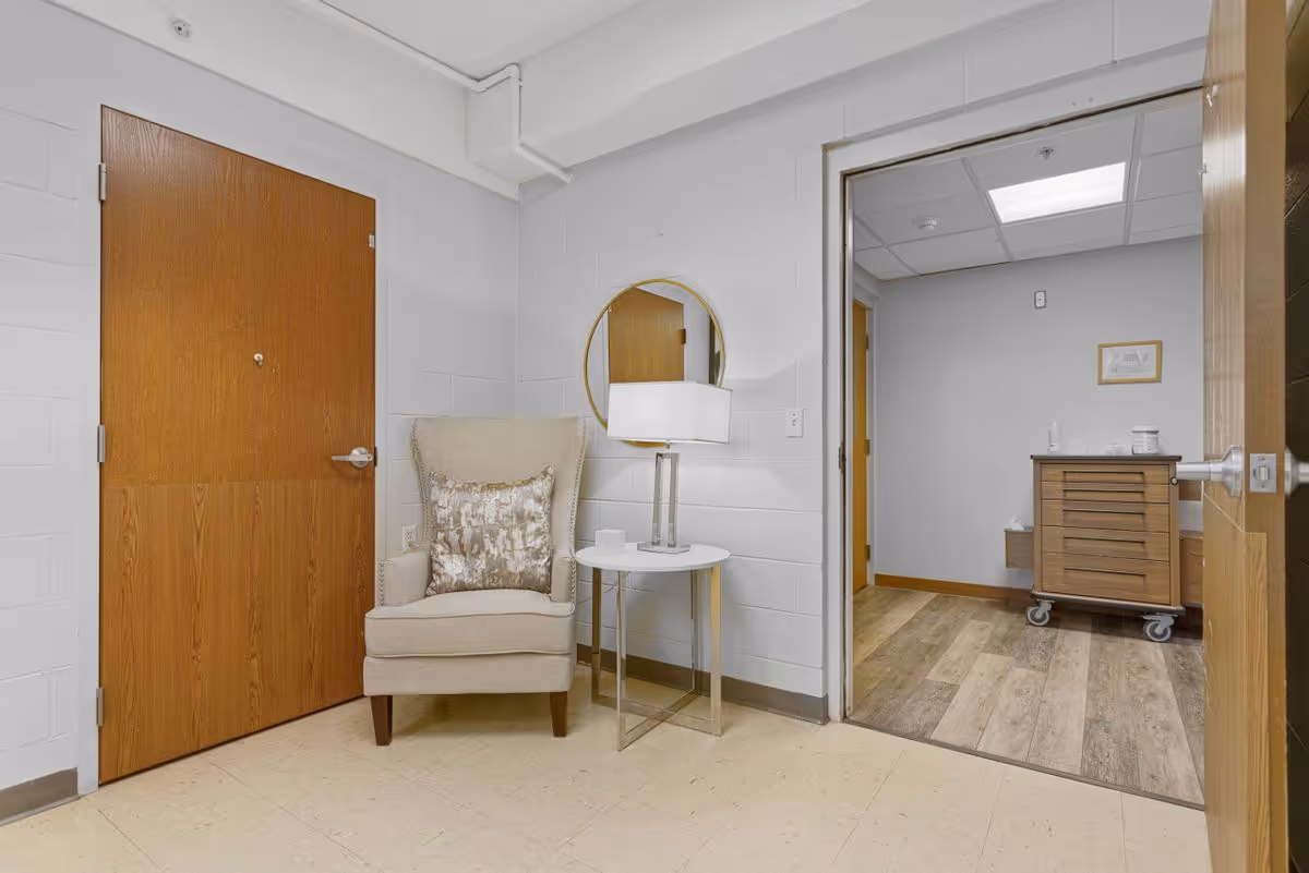 A small interior sitting area with an armchair, side table and lamp beside a wooden door and an open doorway leading to a room with a wheeled cabinet.
