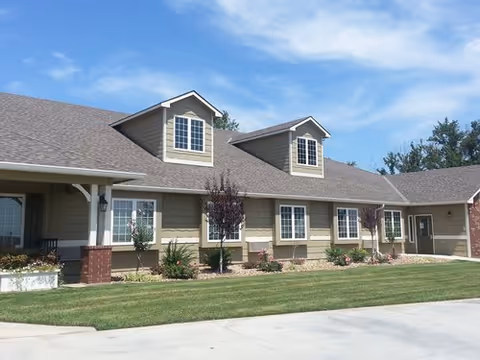 Exterior view of a single-story residential building with beige siding, white trim, and a gray shingled roof under a blue sky with some clouds. The building has multiple windows and small dormers, a covered porch area, landscaped garden beds with shrubs and flowers, and a well-maintained lawn in front.