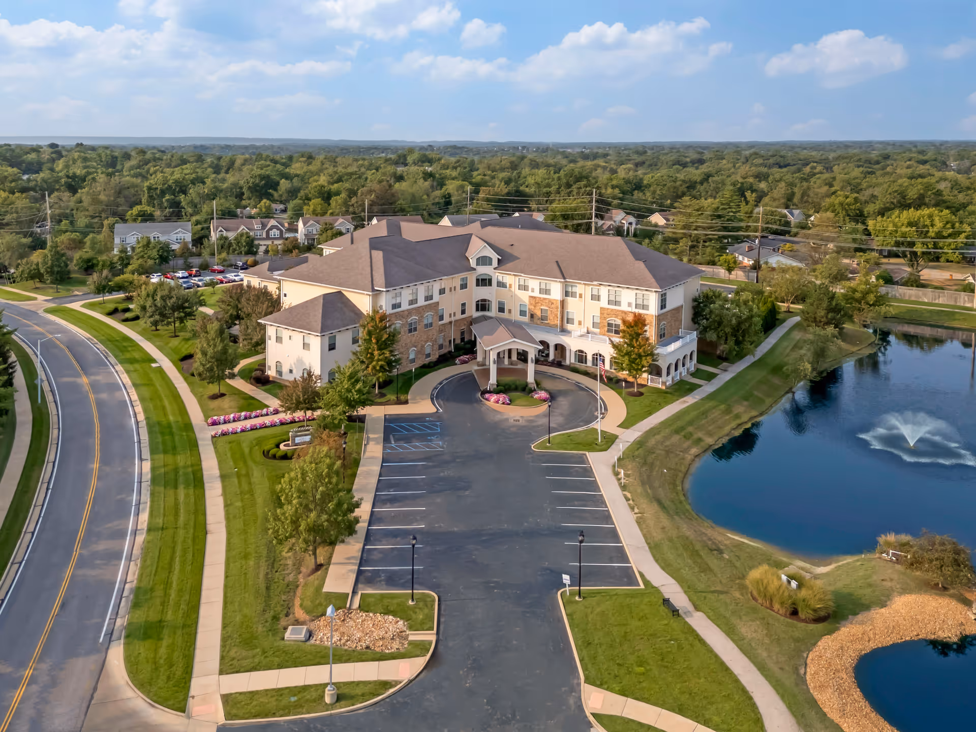 Aerial view of Brookdale West County, a large senior living facility building with a driveway and parking lot in front. The building is surrounded by well-maintained lawns, trees, and flower beds. To the right, there is a pond with a fountain. A road runs along the left side of the property, and residential houses are visible in the background under a partly cloudy sky.
