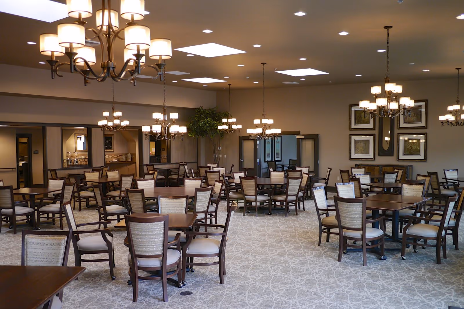 A spacious dining room with multiple wooden tables and cushioned chairs arranged neatly. The room features several elegant chandeliers hanging from the ceiling, recessed lighting, and skylights. There is a large plant in the corner and framed artwork on the walls. The carpet has a subtle patterned design, and there are double doors leading to another room.