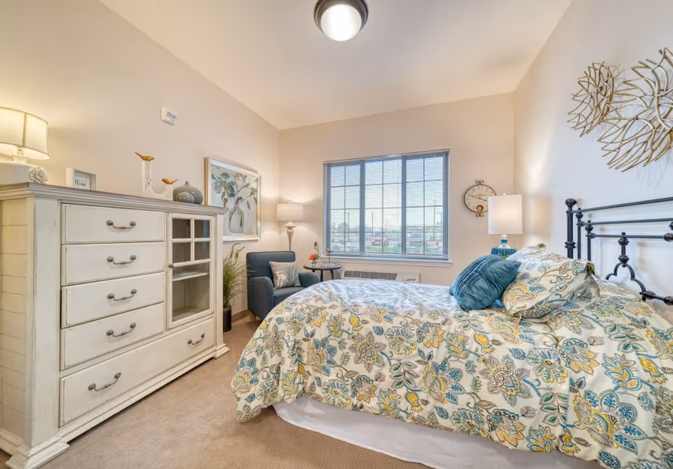 A cozy bedroom in The Woodlake Senior Living facility featuring a bed with floral patterned bedding and blue accent pillows. There is a white dresser with drawers and glass cabinet doors on the left side, a blue armchair with a small round table beside it near the window, and two lamps providing warm lighting. The walls are decorated with framed artwork and a decorative clock.